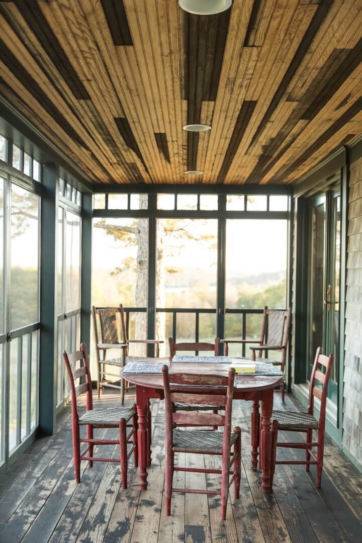 Rustic sunroom with red chairs, wooden table, and large windows overlooking nature