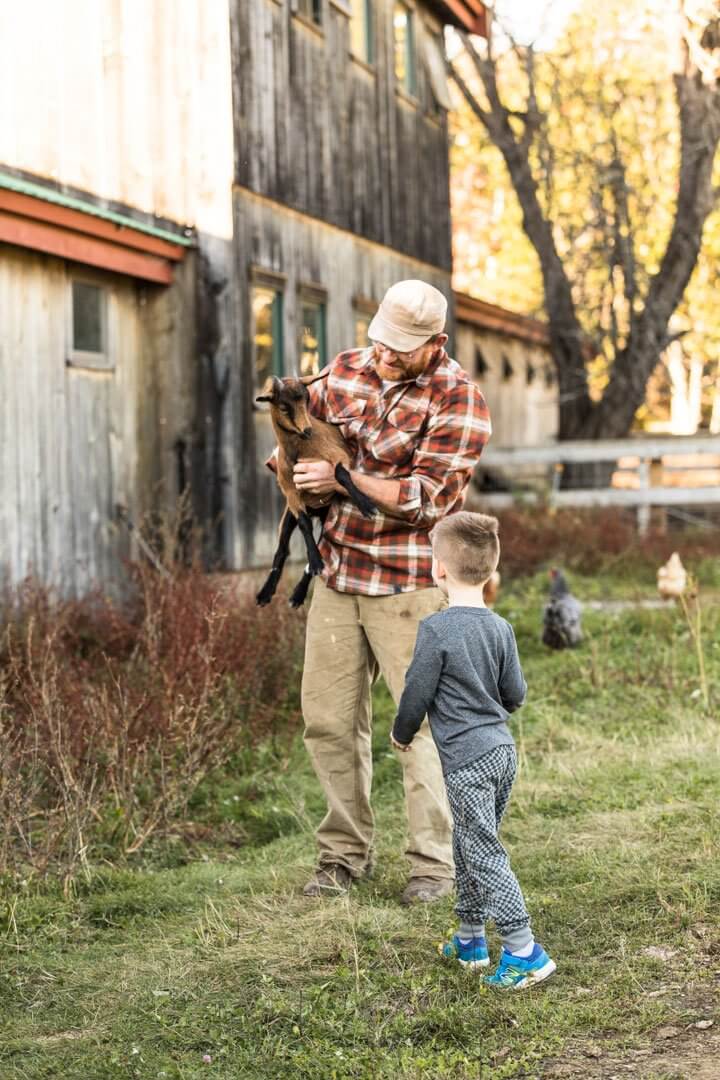 Adult holds baby goat while child watches near rustic barn in autumn