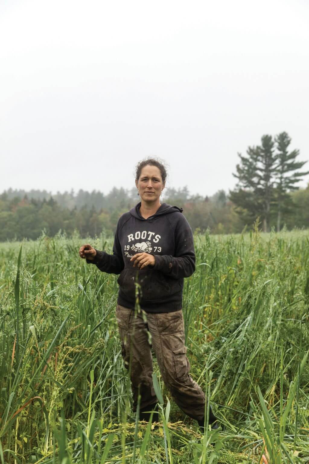 Farmer in Roots hoodie standing in lush green field with forest background