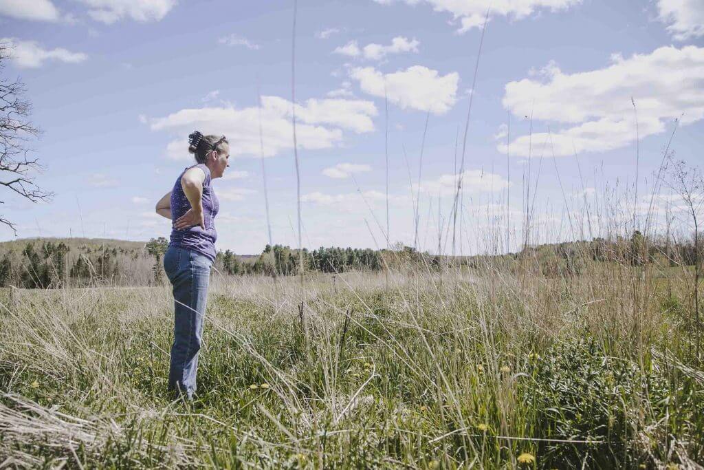 Person standing in tall grass field looking out at cloudy sky and distant trees