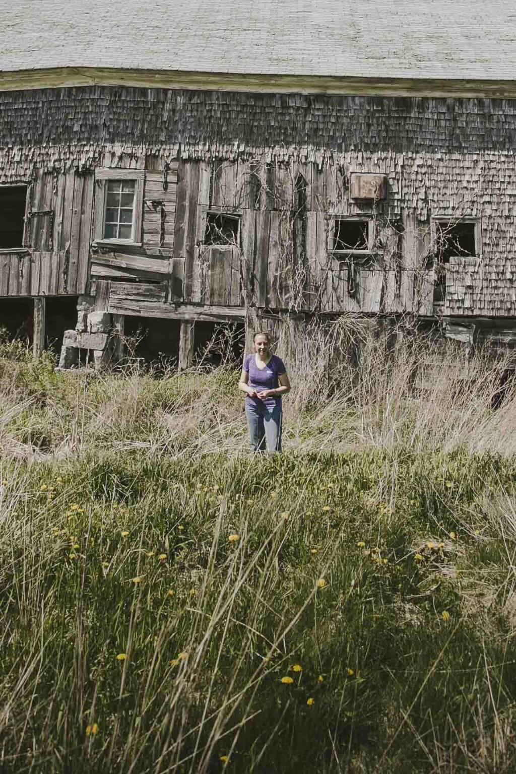 Person standing in front of an old, weathered wooden barn surrounded by tall grass