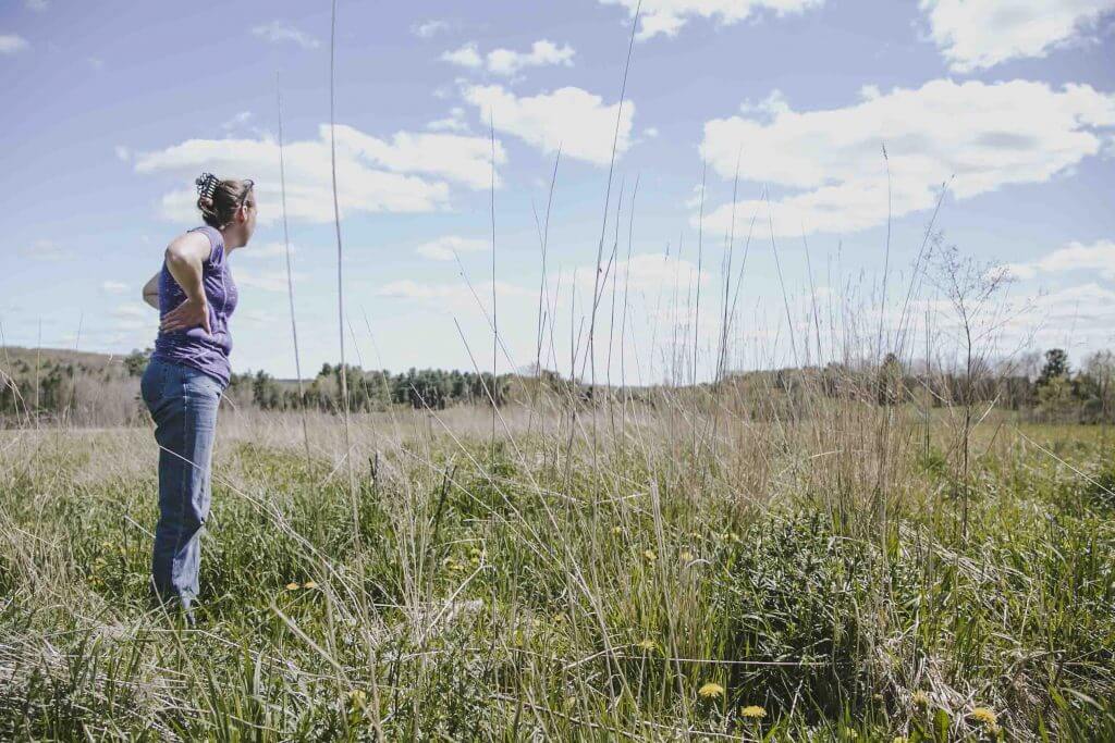 Person standing in grassy field looking towards distant trees and cloudy sky