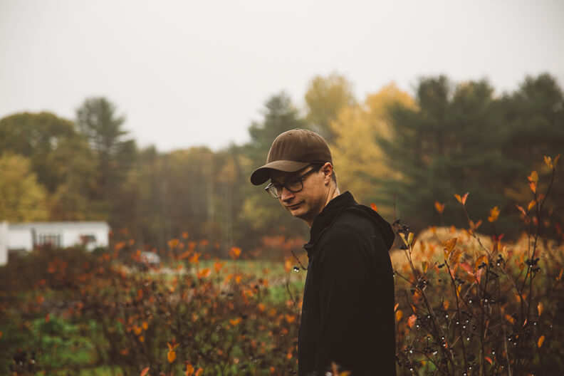 Person in black jacket and cap standing in autumn field with colorful trees