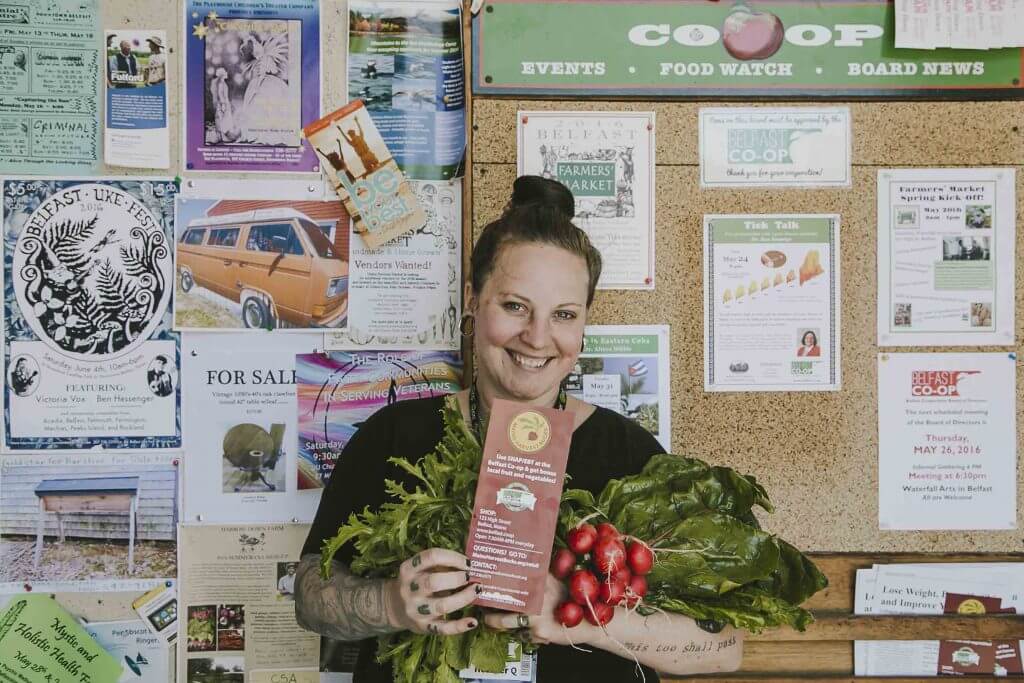 Smiling woman holds fresh radishes and greens in front of community bulletin board