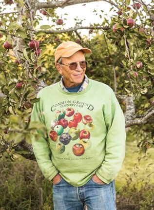 Smiling older person in apple orchard wearing green sweatshirt with fruit design