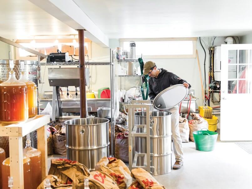Brewery worker operating equipment among large metal brewing tanks and supplies