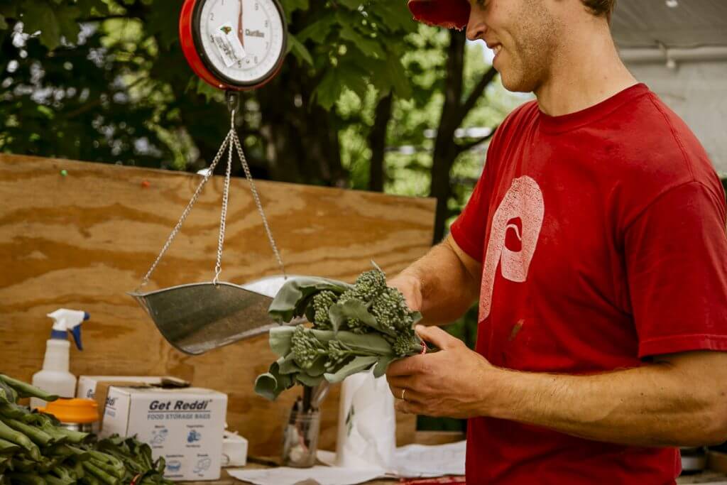 Person in red shirt weighing fresh broccoli on hanging scale outdoors