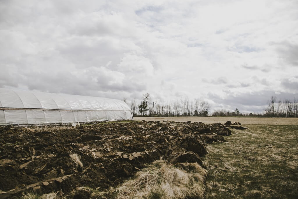 Large white greenhouse in rural field with plowed earth under cloudy sky