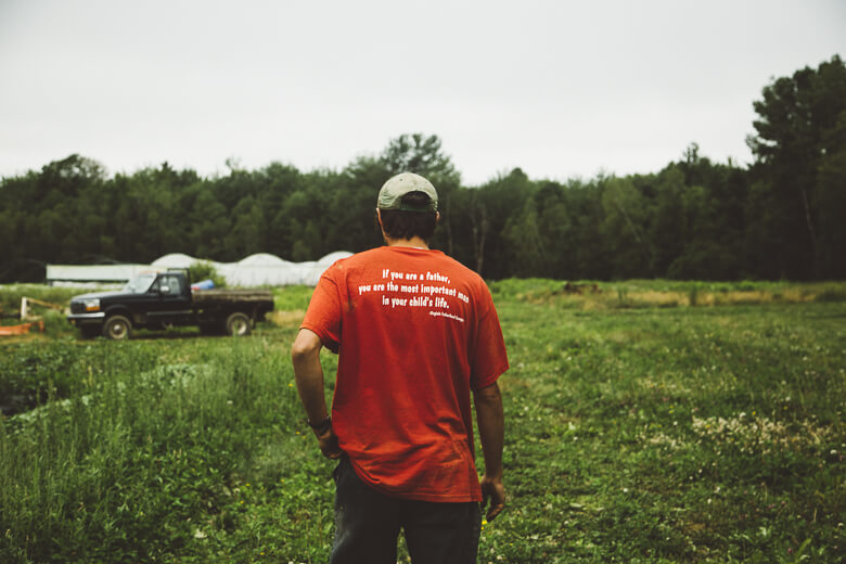 Person in red shirt standing in grassy field with truck and trees