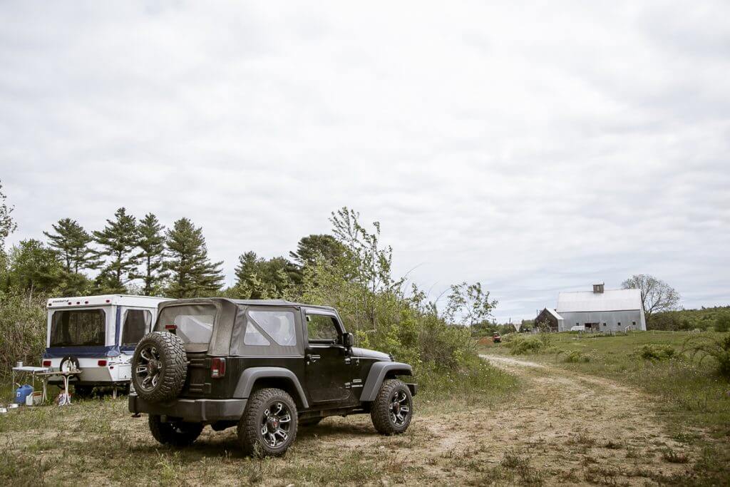 Jeep Wrangler parked with trailer near rural barn and pine trees