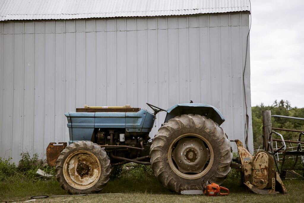 Blue vintage tractor parked beside metal barn with chainsaw in foreground