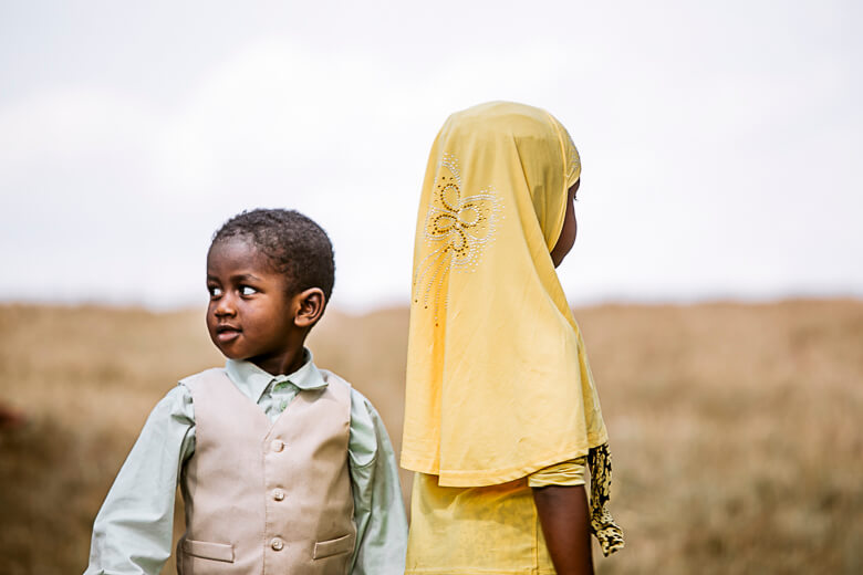 Two children standing together in a field, one in yellow hijab, one in vest