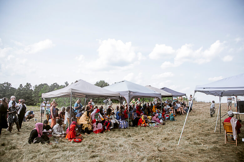 Large community gathering under white tents on grassy field with diverse people