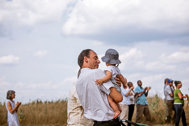 Father holding young child in rural field with people in background