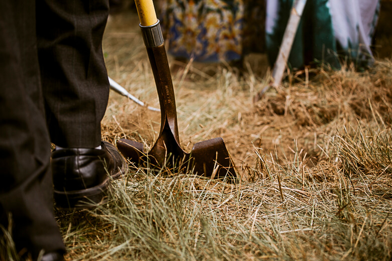 Shovel digging into dry grass and soil with person's legs visible