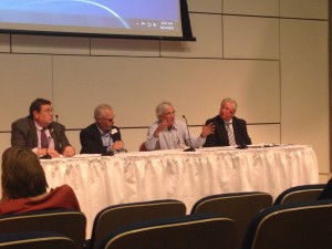 Four professionals seated at a conference panel table with presentation screen