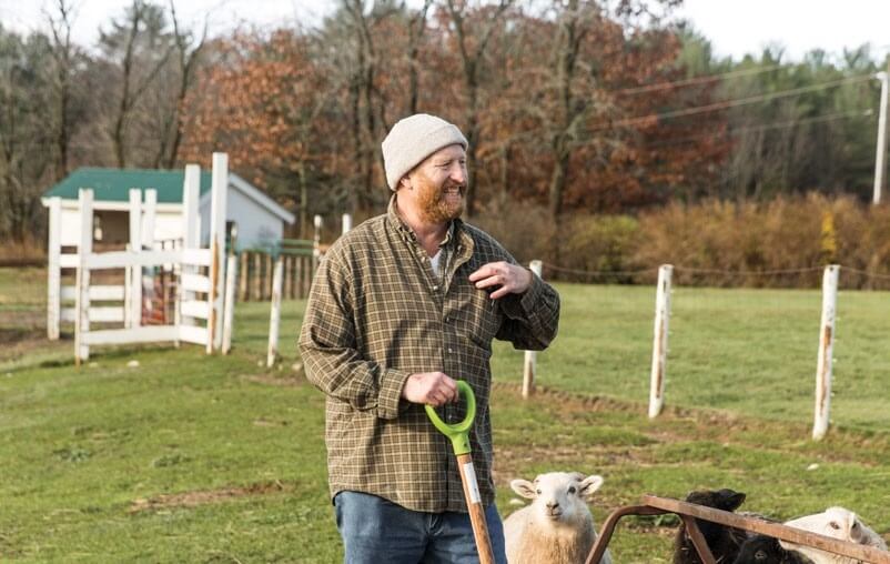 Farmer in beanie with shovel standing near sheep in pasture