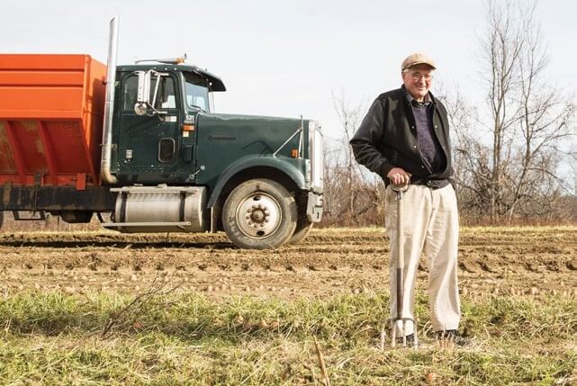 Elderly farmer stands with pitchfork next to large truck in plowed field