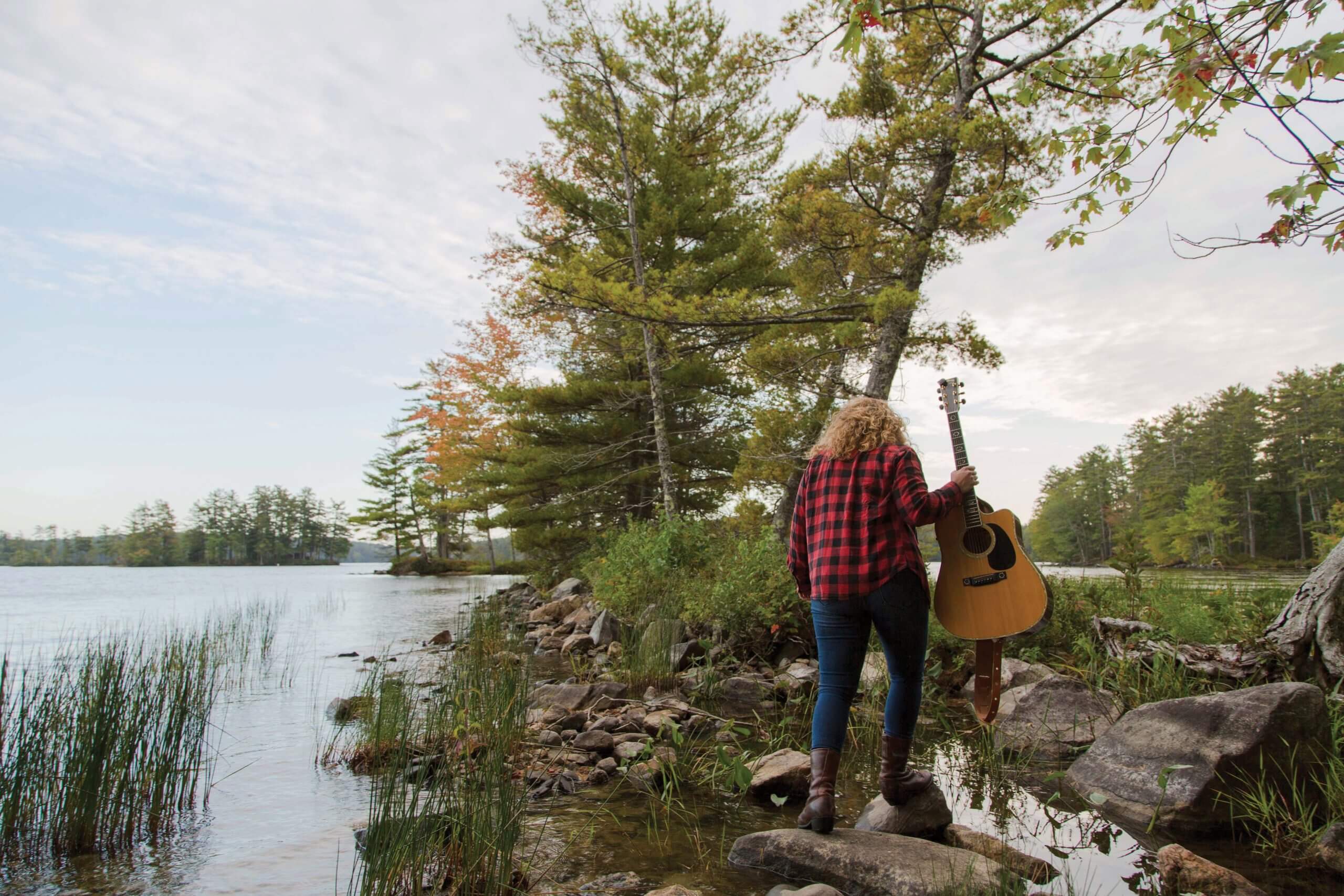 Musician in plaid shirt carrying guitar by rocky lakeside with pine trees