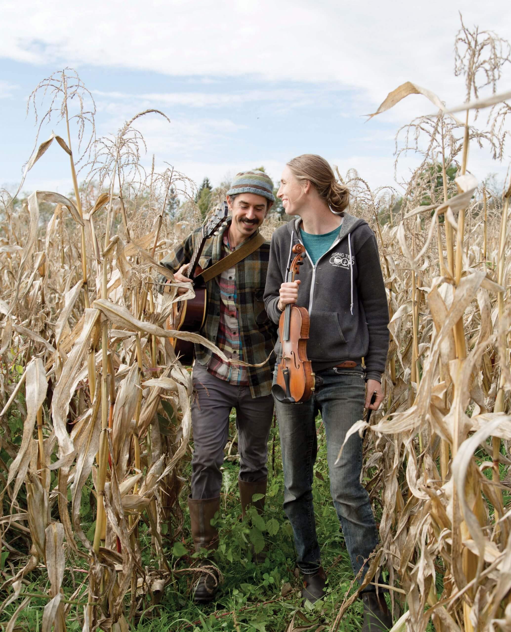 Two musicians walking together through a dry cornfield, carrying guitar and violin