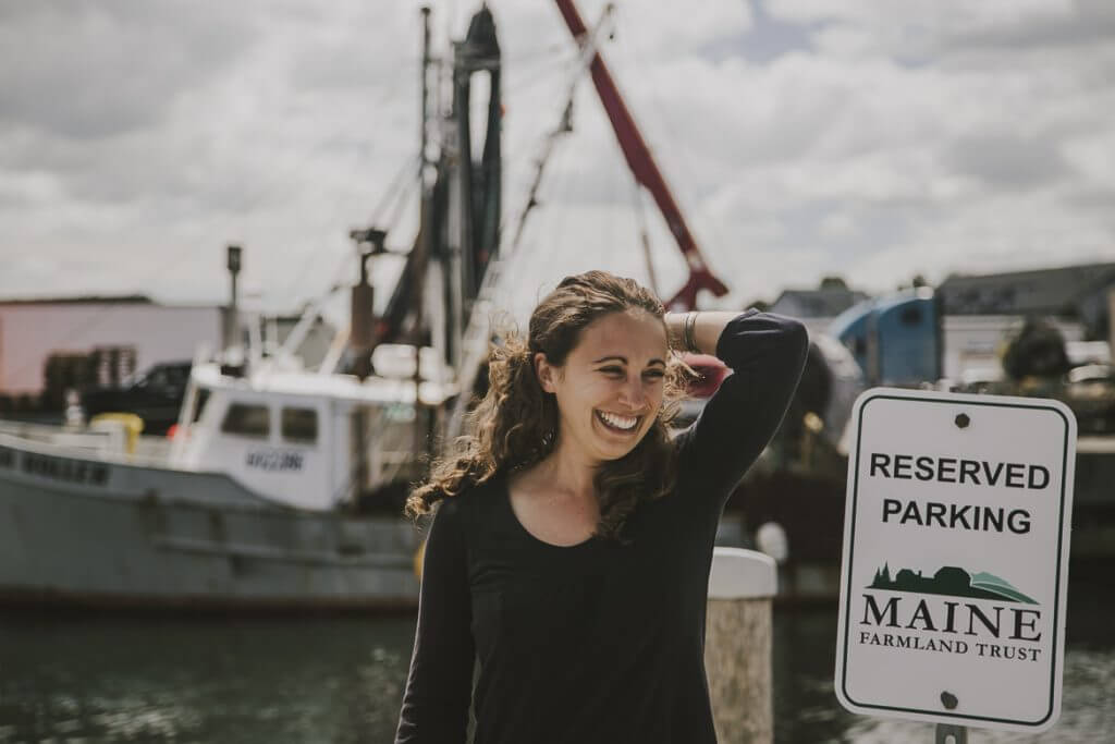 Smiling woman near fishing boats with Maine Farmland Trust parking sign
