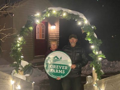 Two people stand under lit greenery arch with Forever Farms sign at night