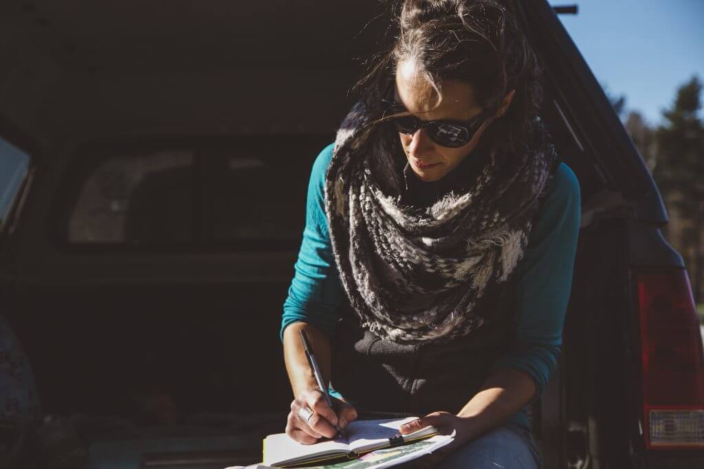 Person in sunglasses writing in notebook near car on sunny day