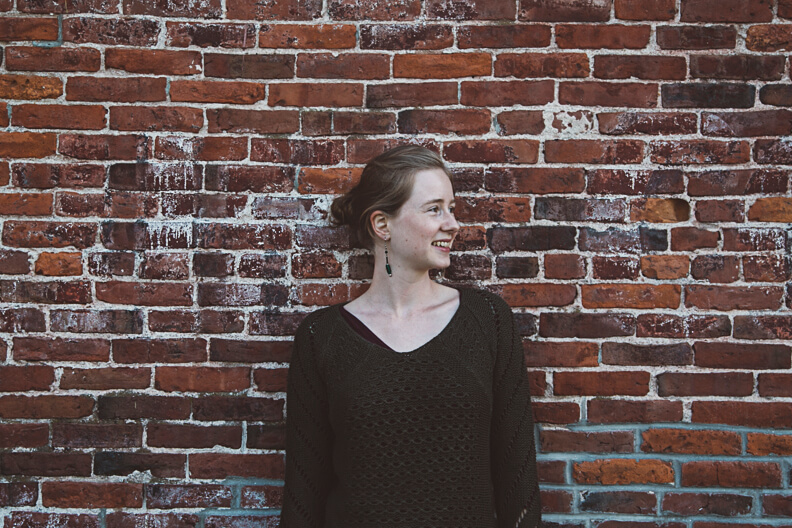 Smiling person in dark sweater standing against weathered brick wall