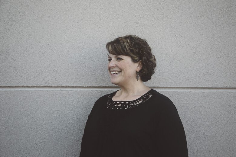 Woman with curly hair in black top laughing against gray wall