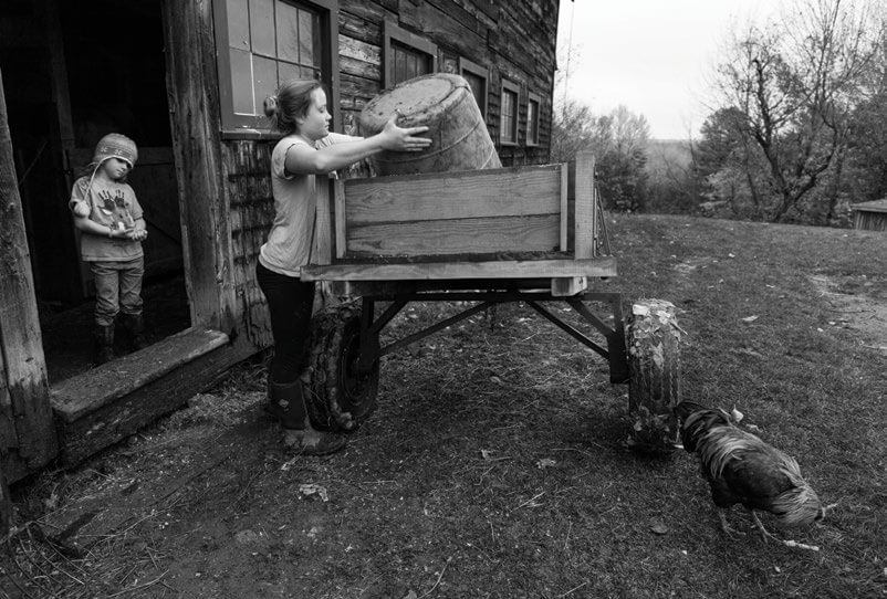 Child lifting heavy bucket on wooden cart near barn, another child watching