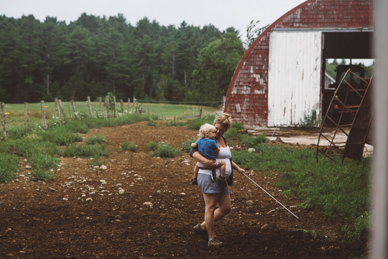 Woman carrying child walks through garden near red barn on farm