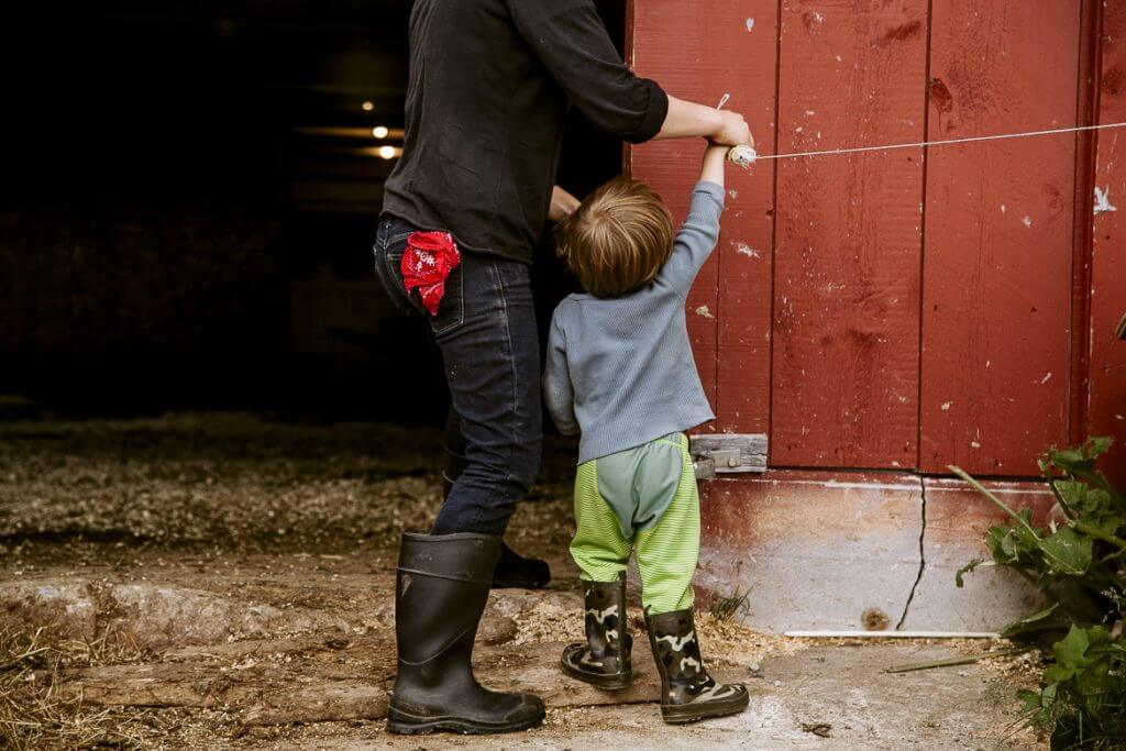 Child in green pants helps adult by red barn with string