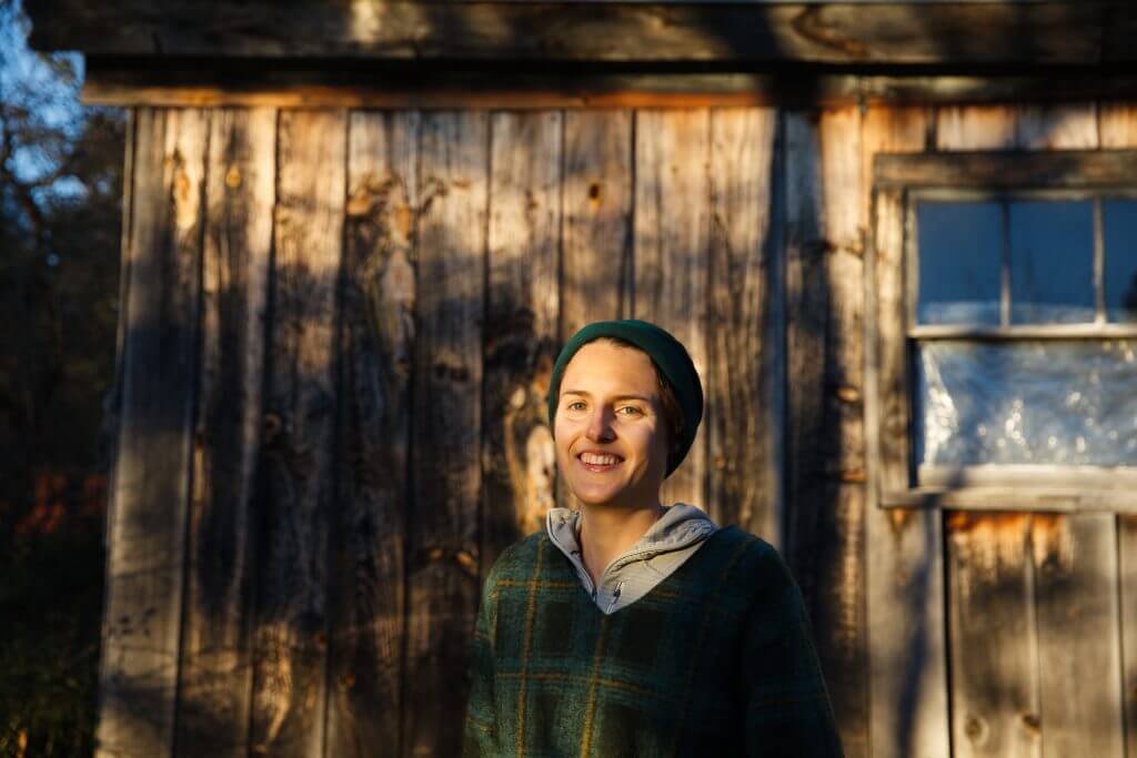 Smiling person in green beanie standing in front of wooden cabin