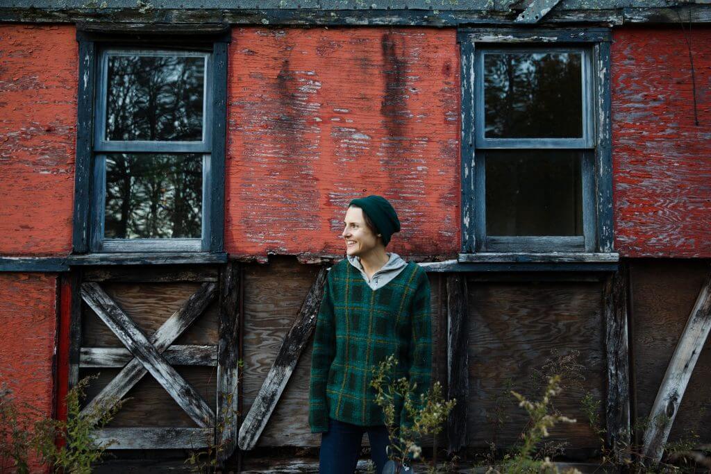Smiling person in green plaid jacket standing by weathered red wooden building