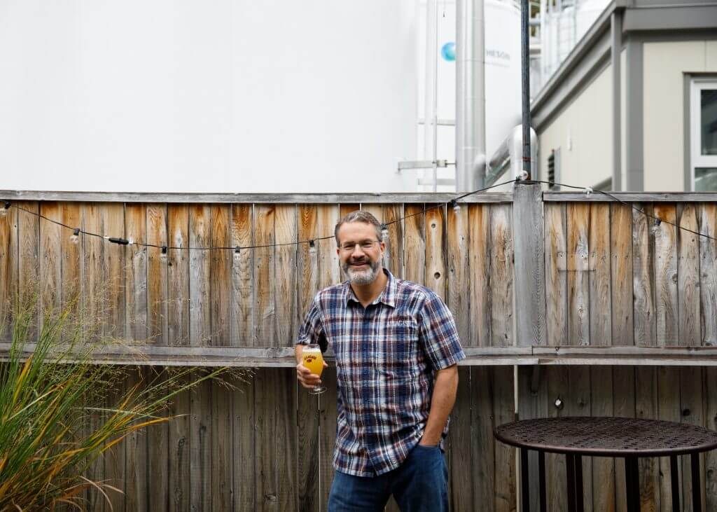 Smiling middle-aged man holding beer in backyard with wooden fence
