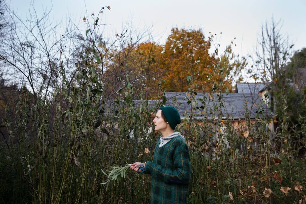 Person in green plaid coat and beanie standing in overgrown autumn garden
