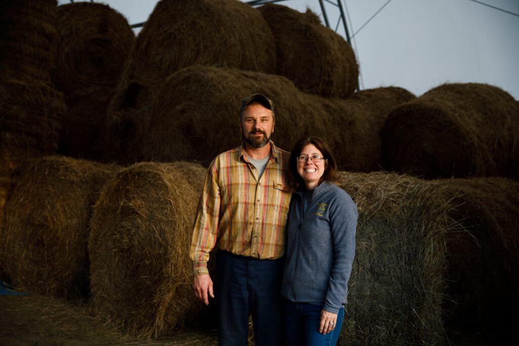 Two farmers standing together in front of large hay bales