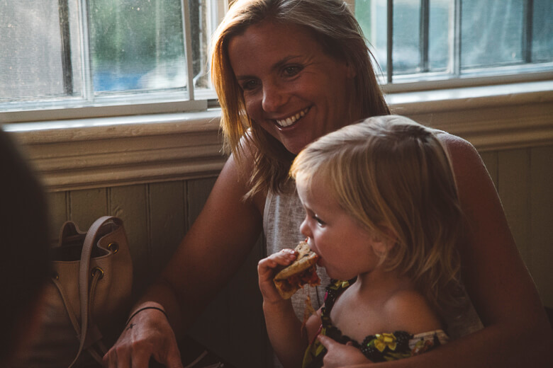 Smiling mother watching young child eat sandwich by window