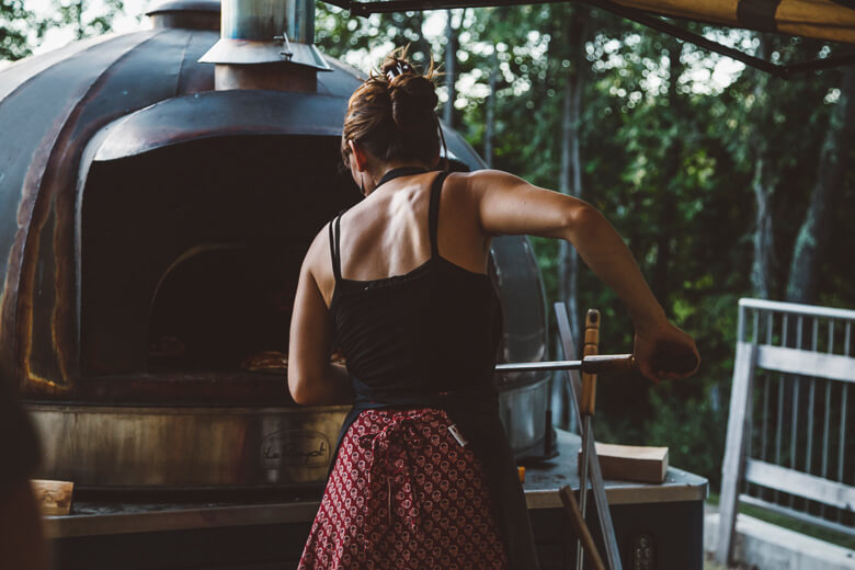 Person cooking at outdoor wood-fired oven surrounded by green trees