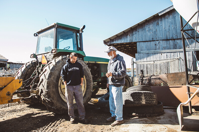 Two workers standing near large John Deere tractor by wooden barn