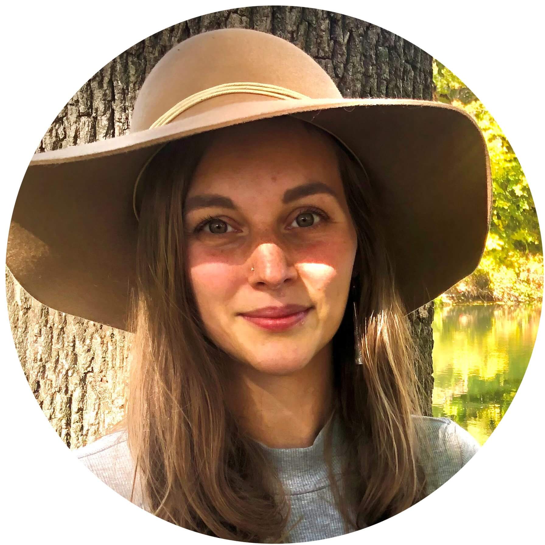 Woman wearing wide-brimmed hat smiling by tree near reflective water