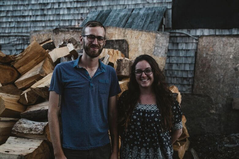 Two people smiling by woodpile in front of weathered wooden building