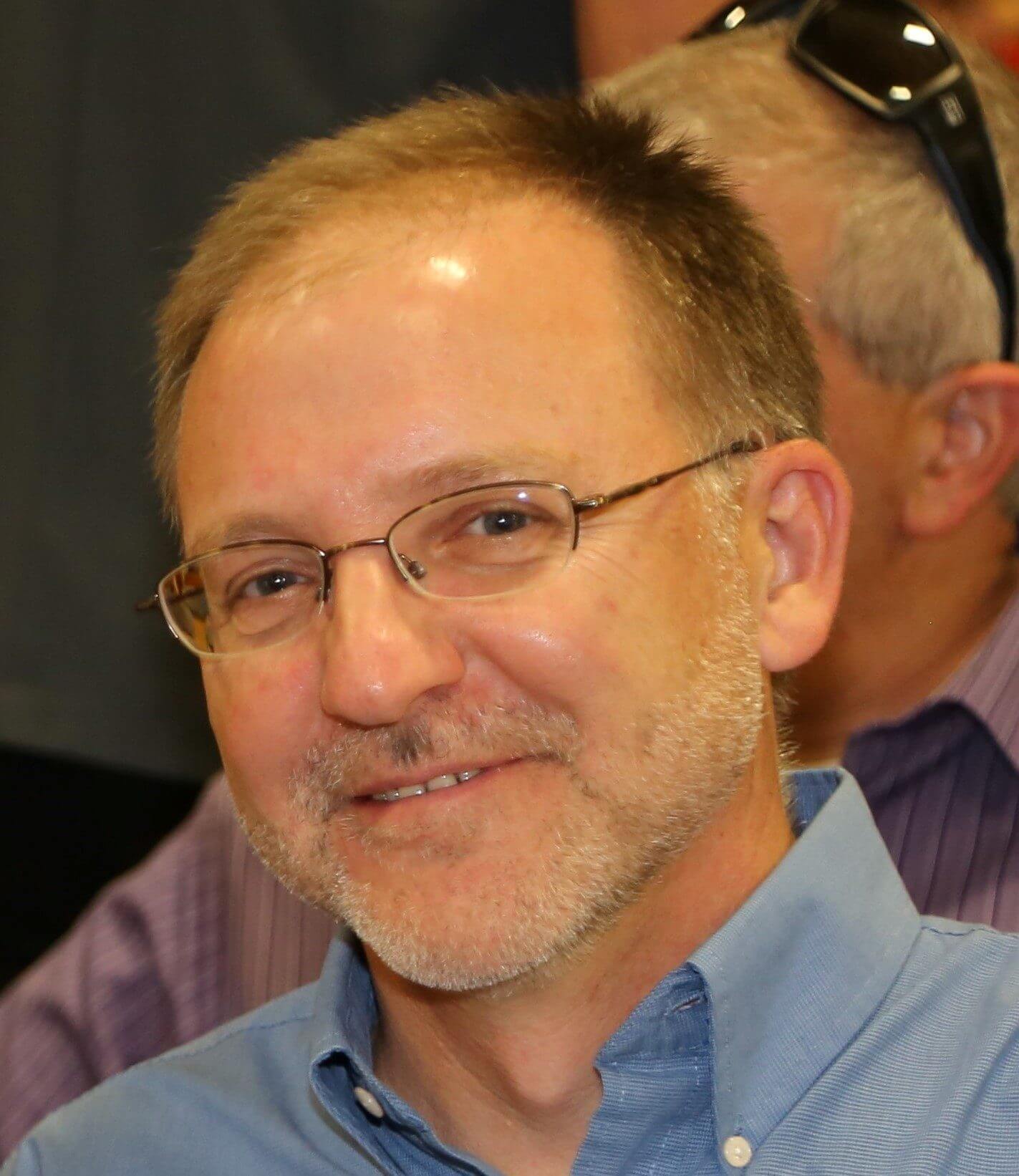Middle-aged man with glasses smiling in blue shirt at camera