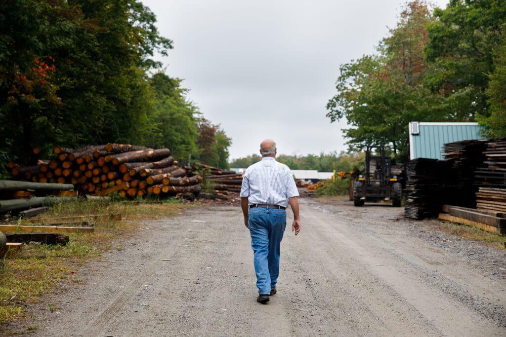 Older worker walking down lumber yard path with stacked logs and equipment