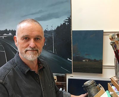 Artist in studio with landscape painting and art supplies