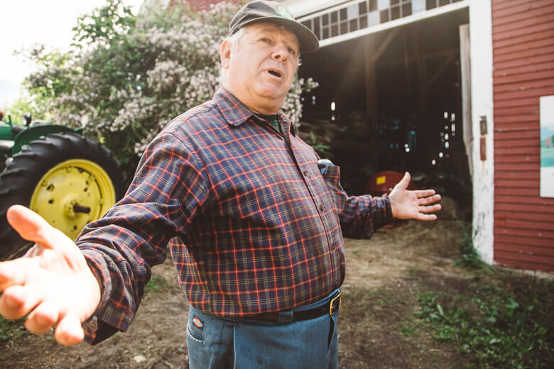 Elderly farmer in plaid shirt gestures near red barn and yellow tractor