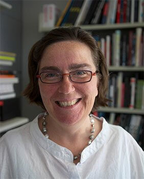 Smiling person in glasses standing in front of bookshelf in white shirt