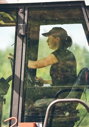 Man in cap operating farm tractor with hands on steering wheel and window visible