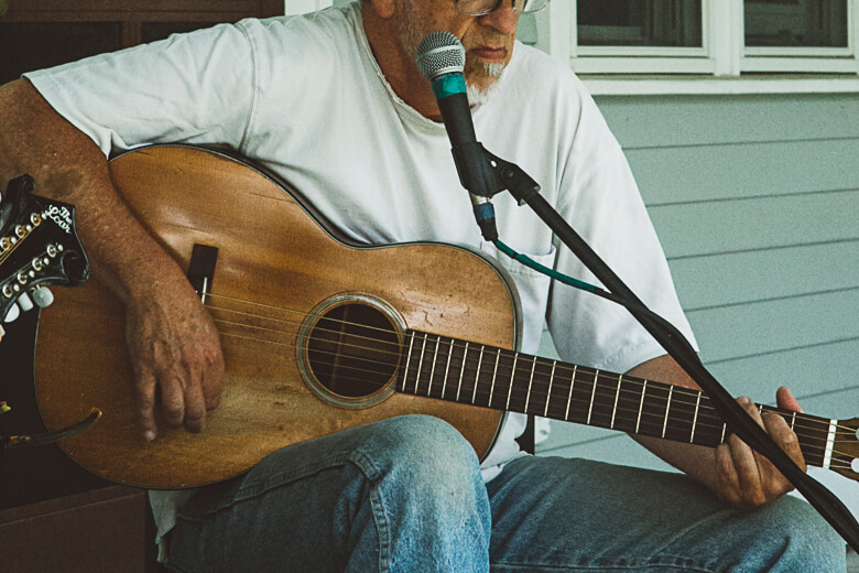 Elderly musician playing acoustic guitar with microphone on porch