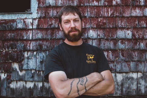Bearded man with crossed arms standing in front of weathered wooden wall